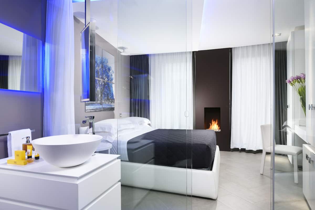 Une chambre à coucher moderne avec un lit entouré de parois de verre, une table de chevet blanche avec des articles de toilette, et une cheminée en arrière-plan.