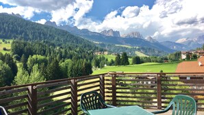 A balcony with a view of a mountainous landscape, including green fields and distant peaks.