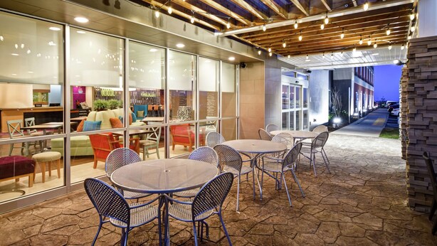 An outdoor seating area with tables and chairs, a glass wall with interior furnishings, and a wooden ceiling with exposed beams and string lights. - Home2 Suites by Hilton Smyrna Nashville (Smyrna)