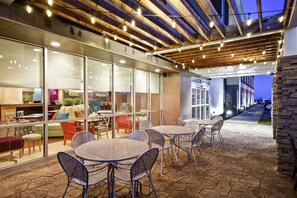 An outdoor seating area with tables and chairs, a glass wall with interior furnishings, and a wooden ceiling with exposed beams and string lights. - Home2 Suites by Hilton Smyrna Nashville (Smyrna)