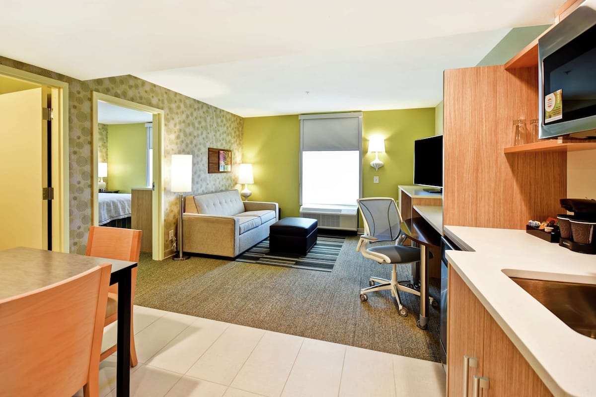 A hotel room with a sofa, a dining table with chairs, a TV, and a kitchenette.