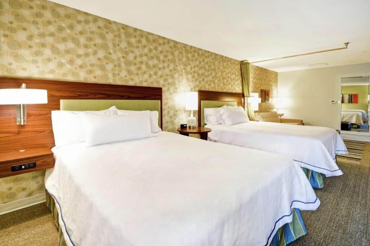 A hotel room with two beds, a wooden headboard, a nightstand, a lamp, and a carpeted floor.