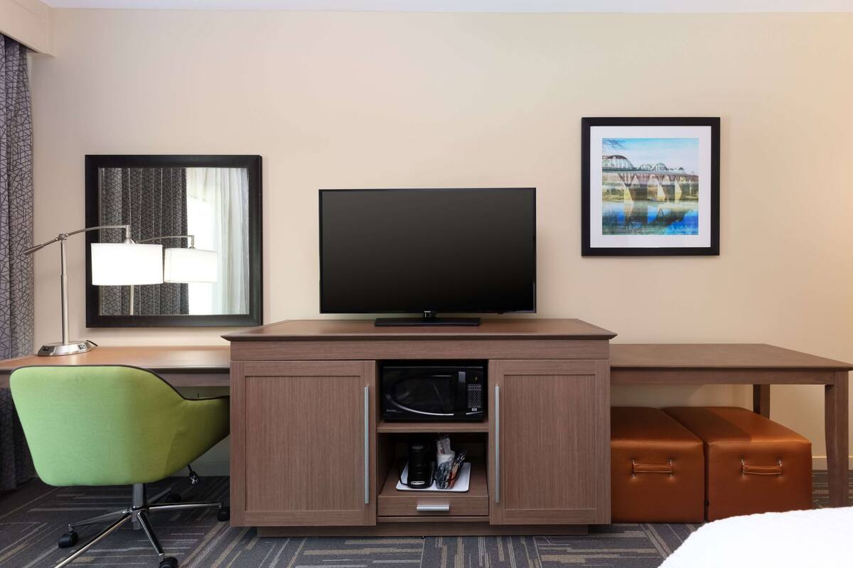 A hotel room with a flat-screen TV, a desk with a microwave, a green chair, and a brown ottoman.