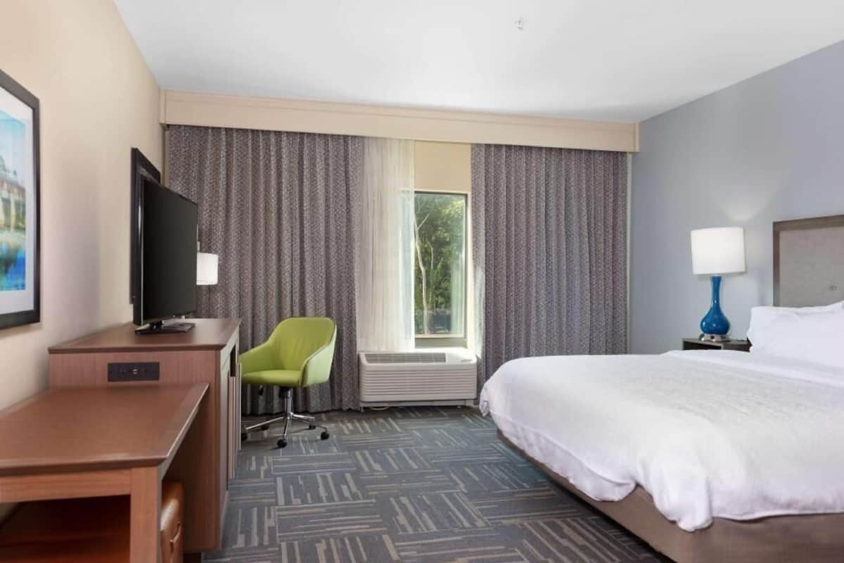 A hotel room with a bed, a desk, a television, a chair, a window with curtains, and a nightstand with a lamp.