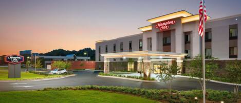 Un hotel Hampton Inn con una entrada bien iluminada, un estacionamiento y una zona ajardinada con árboles.