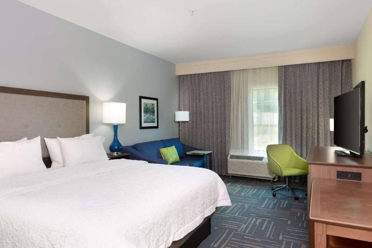 Quarto de hotel com uma cama grande, uma escrivaninha com televisão de tela plana, uma cadeira, um sofá e uma janela com cortinas.