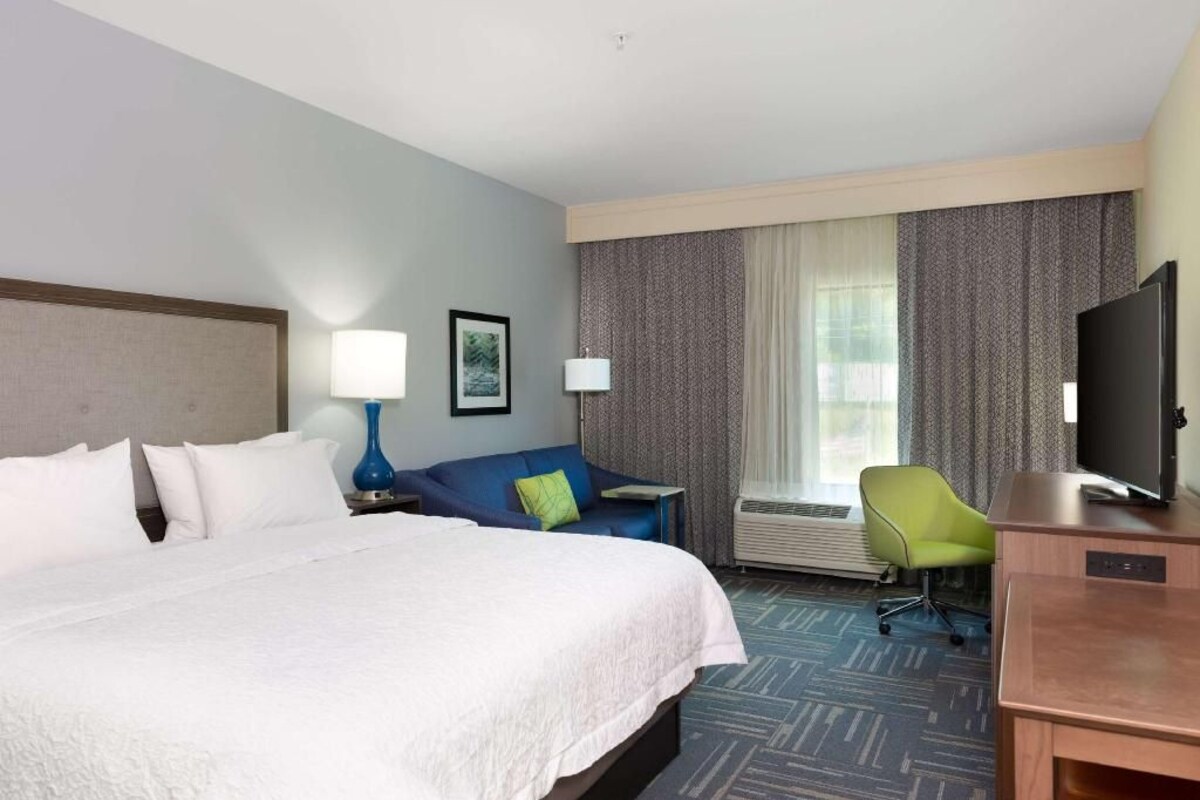 A hotel room with a large bed, a desk with a flat-screen TV, a chair, a sofa, and a window with curtains.