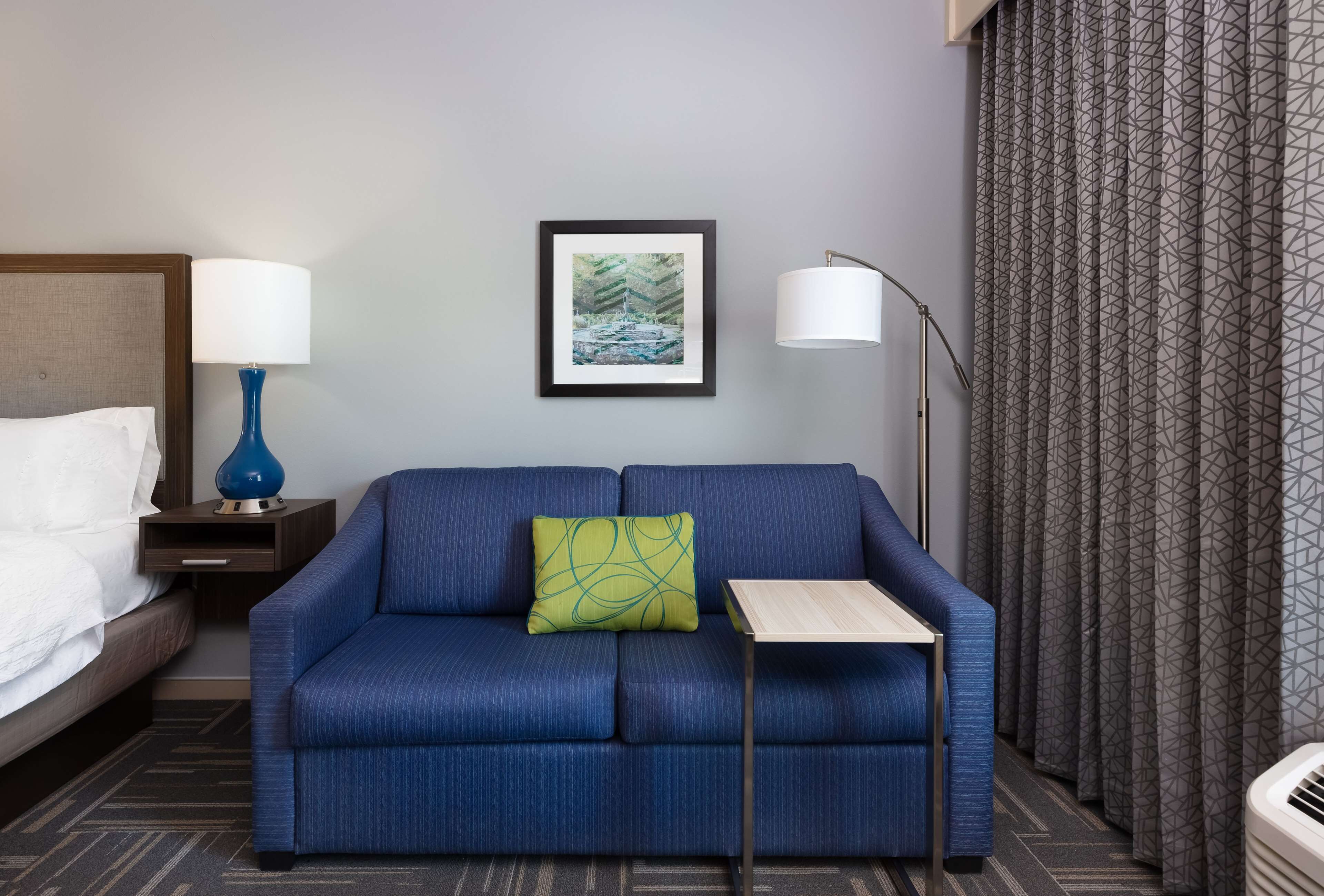 Habitación de hotel con un sofá azul, una mesita, un cuadro enmarcado en la pared, una mesita de noche con una lámpara y una cama con ropa blanca.