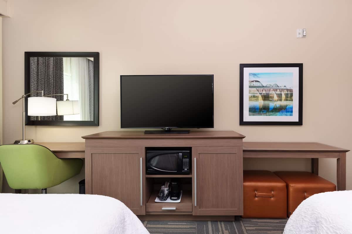 A hotel room with a television, a framed picture on the wall, a desk with a lamp, and a green chair.