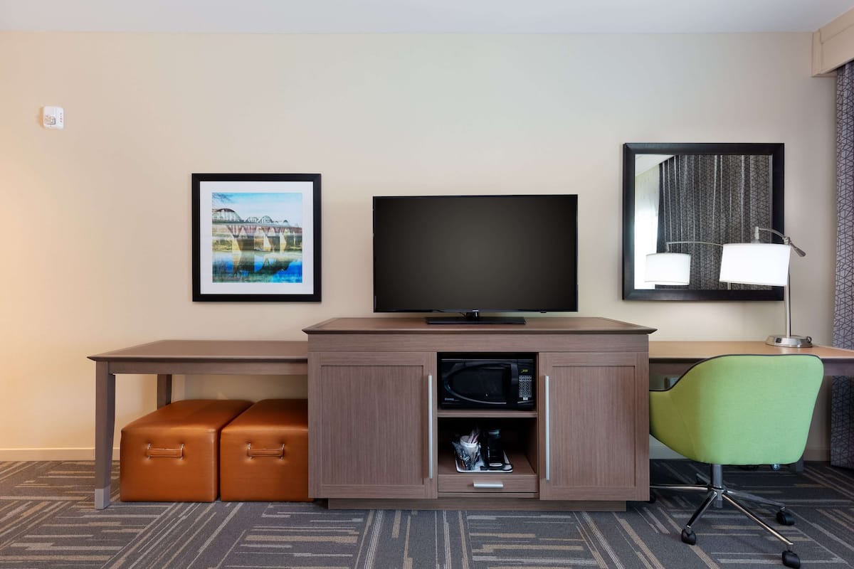 A hotel room with a flat-screen TV, a framed picture, a mirror, a desk, a green chair, and a brown ottoman.