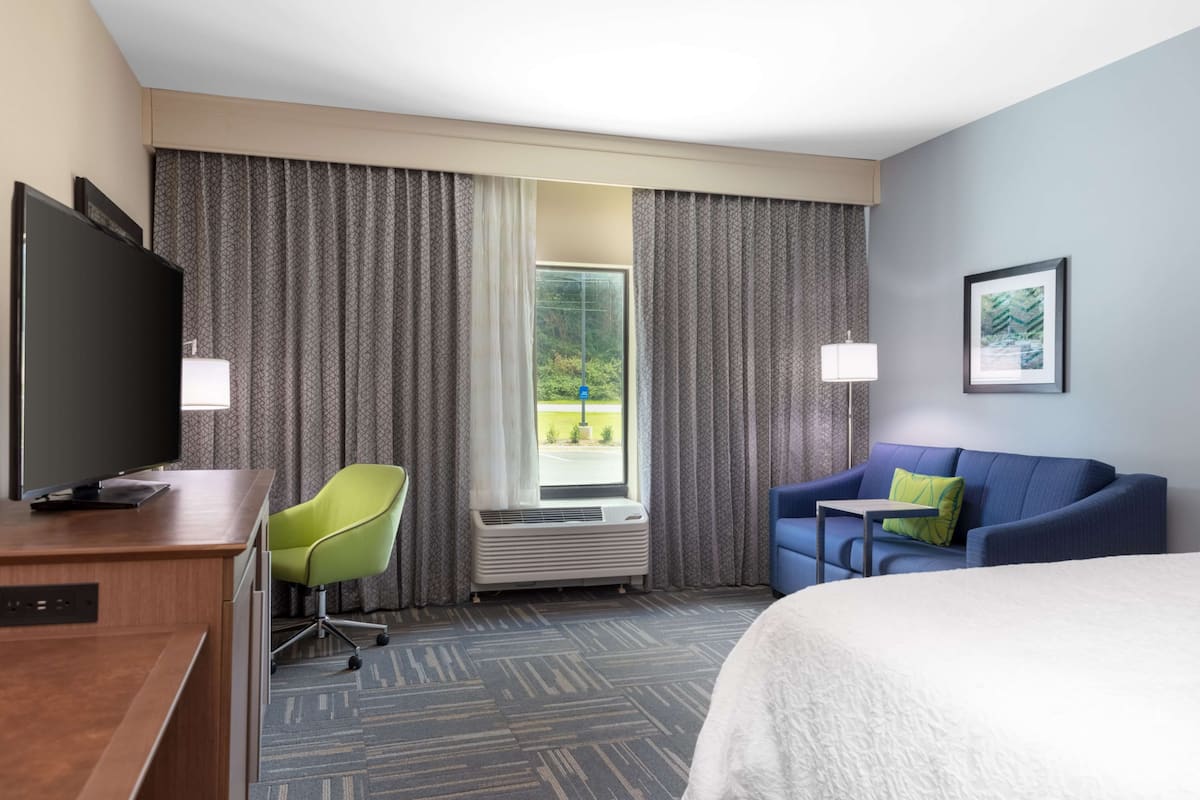A hotel room with a bed, a desk with a TV, a chair, a couch, and a window with curtains.