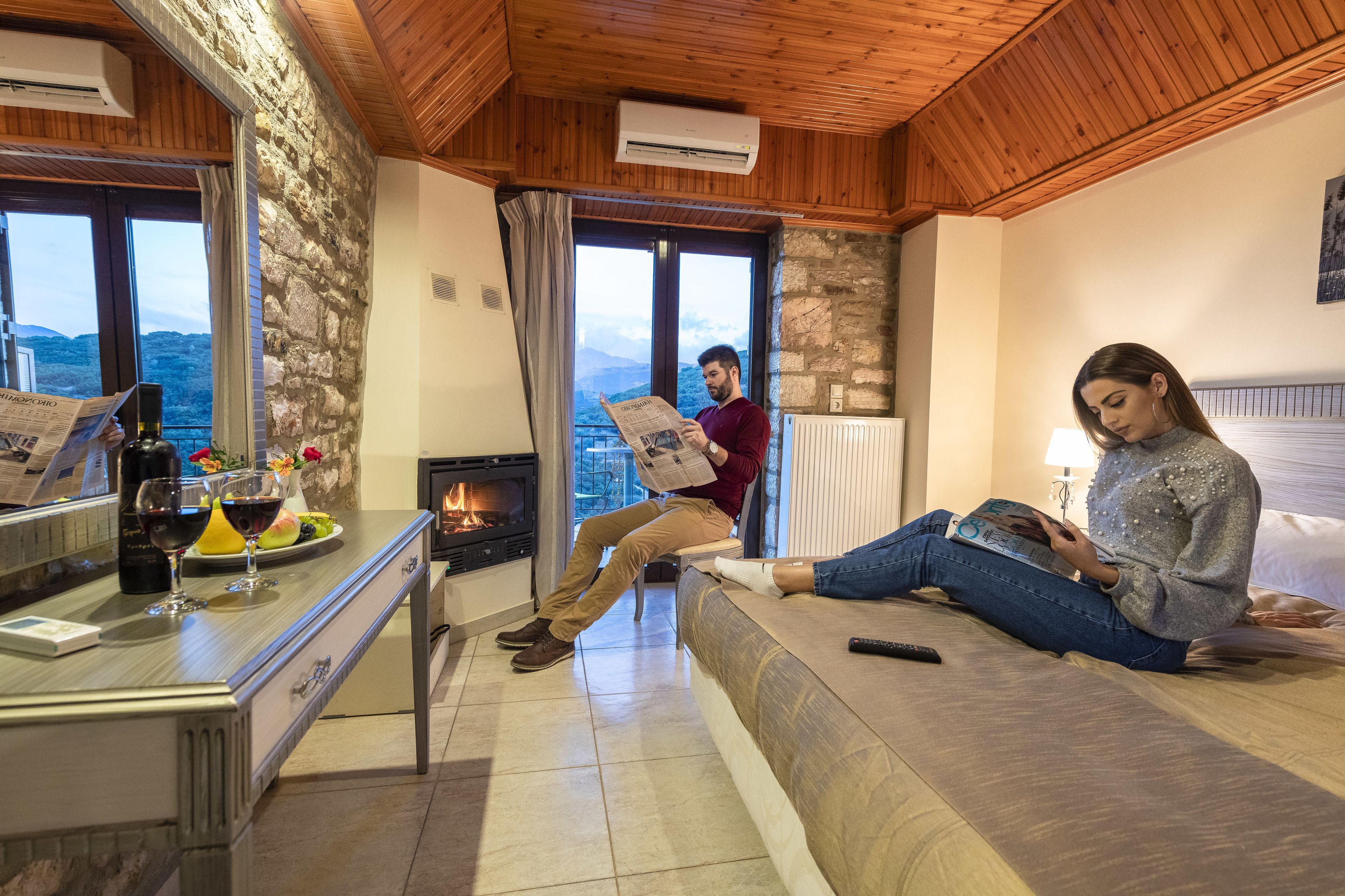 Una habitación acogedora con pared de piedra, techo de madera y chimenea. Una persona está sentada en un sofá leyendo un libro, y otra disfruta de la chimenea.