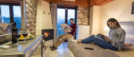 Una habitación acogedora con pared de piedra, techo de madera y chimenea. Una persona está sentada en un sofá leyendo un libro, y otra disfruta de la chimenea.