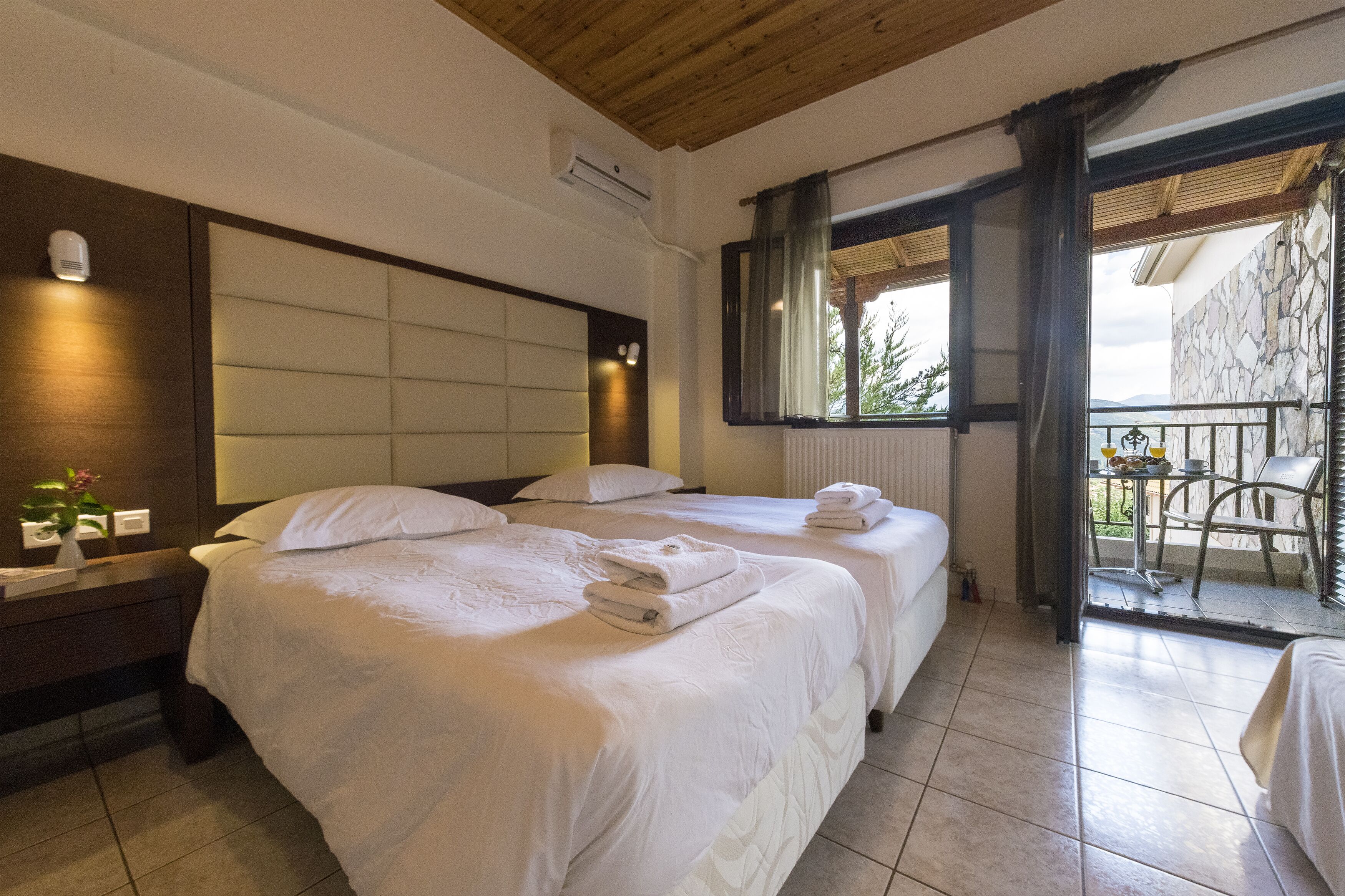 A hotel room with two beds, a wooden ceiling, a balcony with a table and chairs, and a view of the outdoors.
