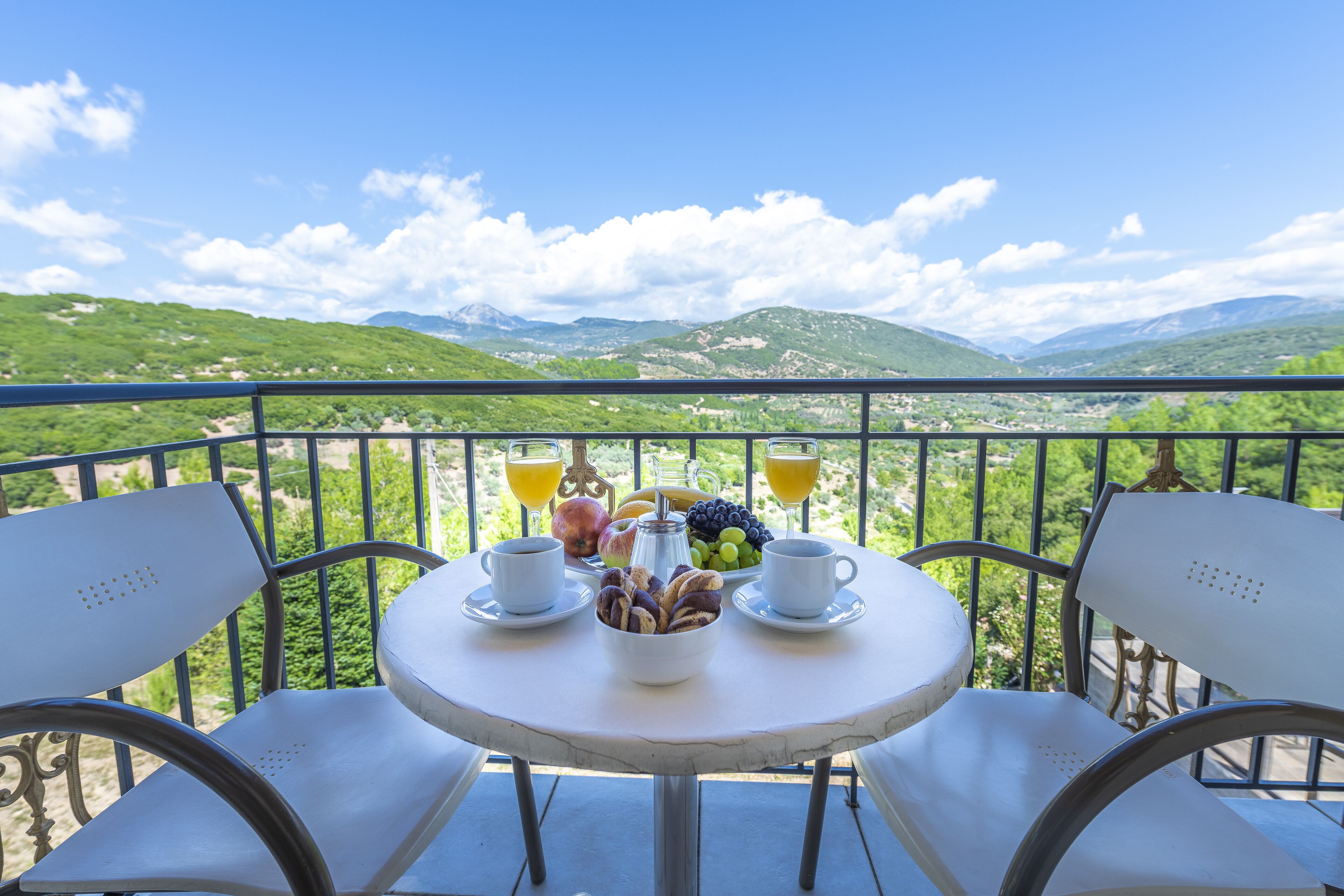 Un tavolo rotondo con sedie bianche, un boccale di vetro con caffè, una ciotola di pasticcini e una coppetta di frutta con uva e una mela su un balcone affacciato su dolci colline e montagne.