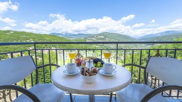 Un tavolo rotondo con sedie bianche, un boccale di vetro con caffè, una ciotola di pasticcini e una coppetta di frutta con uva e una mela su un balcone affacciato su dolci colline e montagne.