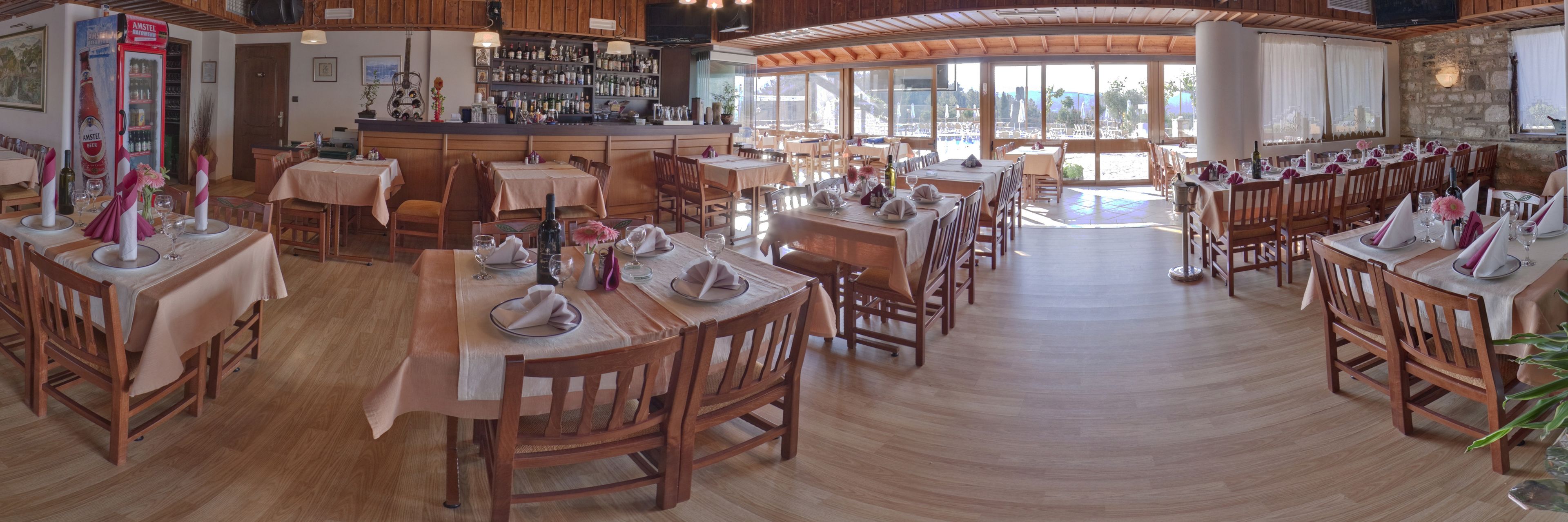 A restaurant with wooden tables and chairs, a bar area, and large windows offering a view of the outdoors.