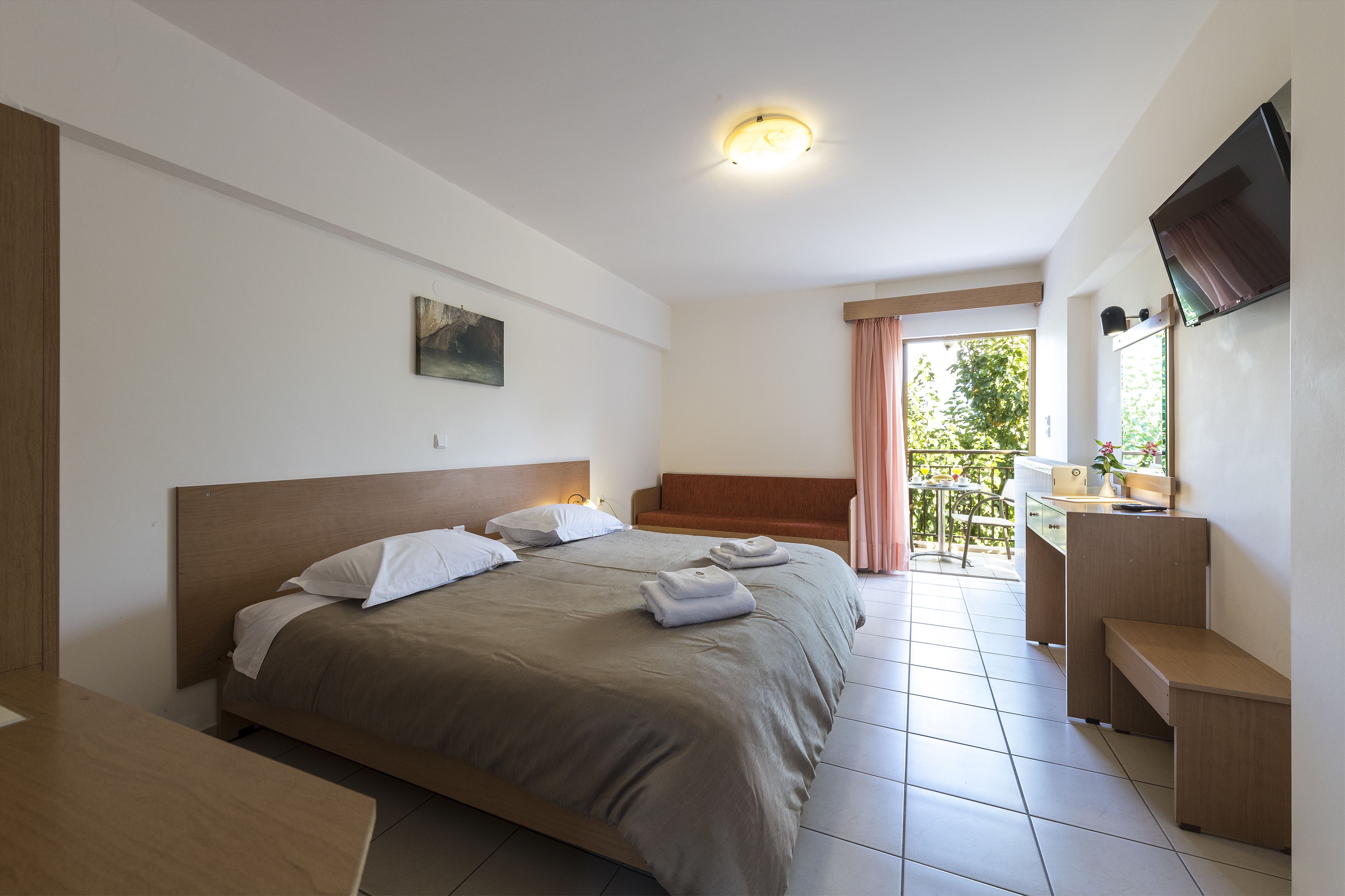 A hotel room with a large bed, a desk, a chair, a TV, and a balcony with a view of greenery.