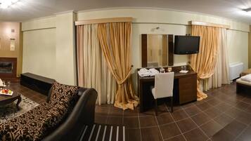 A hotel room with a sofa, a small table with a tray of fruit, a TV, a desk with a mirror, a bed with towels, and a shower area.