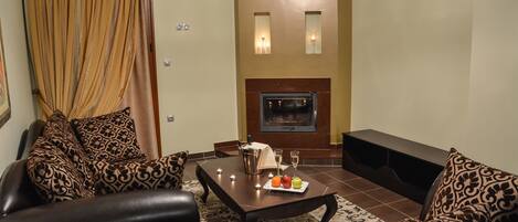 Una sala de estar con chimenea, sofás de cuero, una mesa de centro con frutas y bebidas, y una cortina.