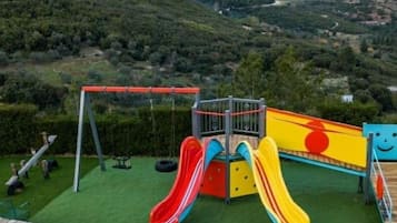 Un parque infantil con toboganes y columpios, con colinas y montañas verdes de fondo.