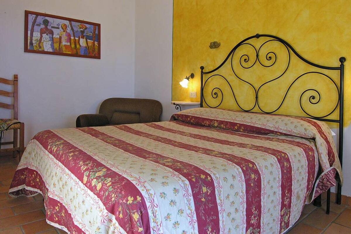 A bedroom with a bed, a chair, a painting on the wall, and a wall-mounted lamp.