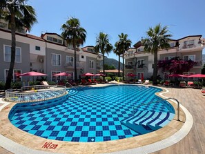 Una piscina con motivo a scacchi blu, circondata da lettini e ombrelloni, con un edificio sullo sfondo.