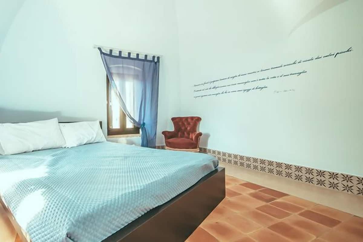 A bedroom with a bed, a nightstand, a chair, a window with curtains, and a wall with a quote.
