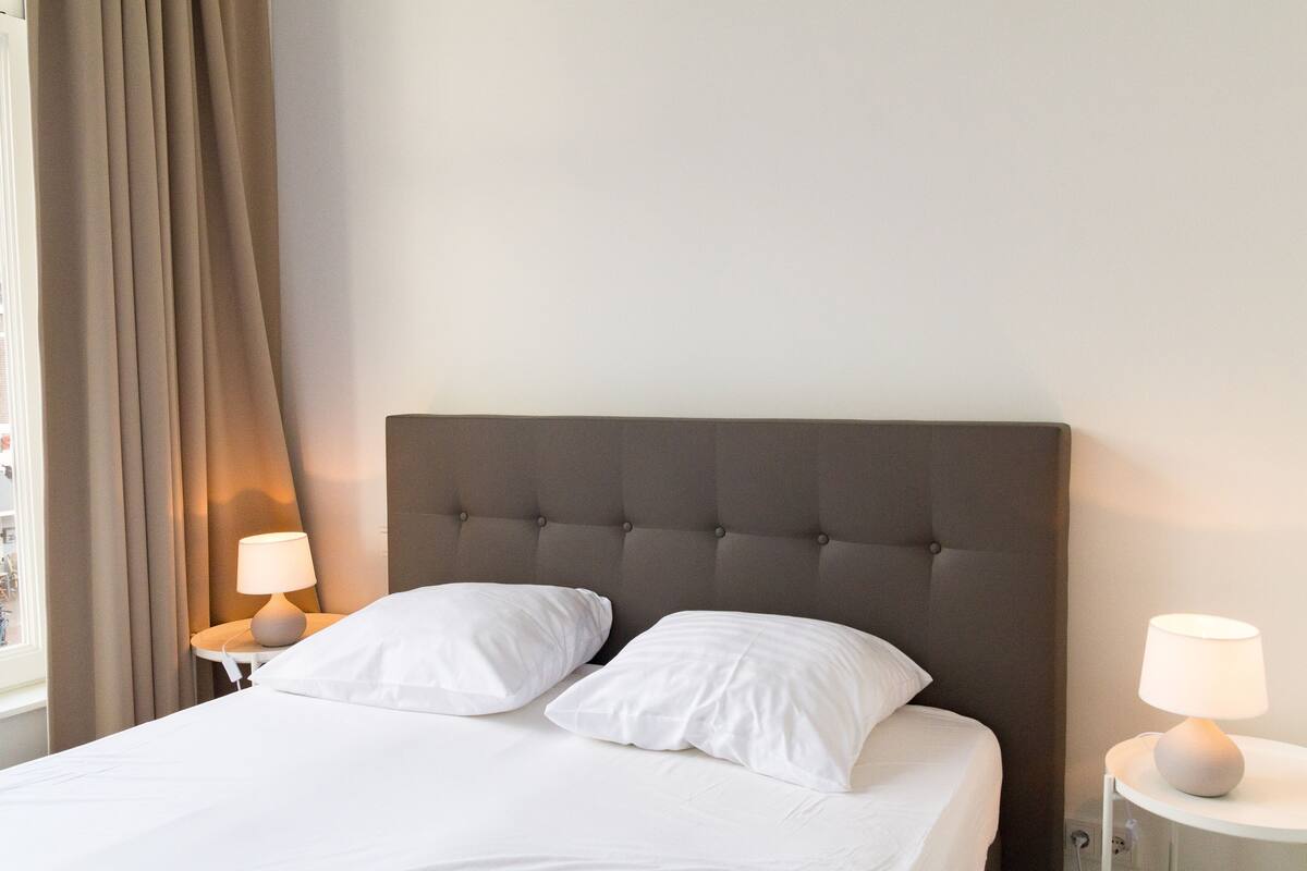 Cama bem arrumada com duas almofadas, cabeceira estofada e duas luminárias de cabeceira em mesas de cabeceira brancas.