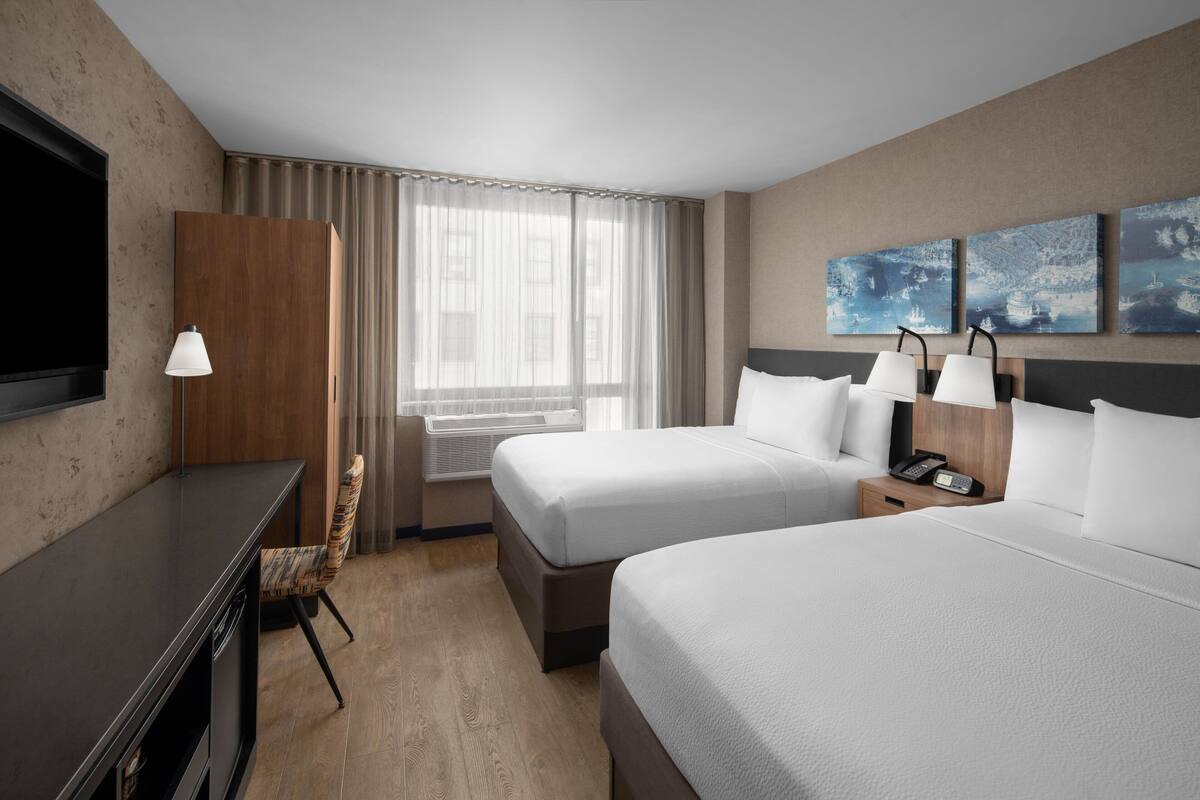 Ein Hotelzimmer mit zwei Betten, einem Schreibtisch, einem Stuhl, einem Fernseher, einem Fenster mit Vorhängen und Bildern an der Wand.