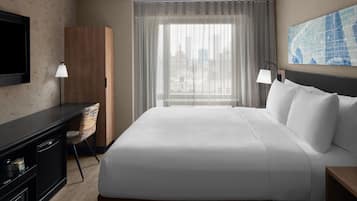 Quarto de hotel com uma cama grande, uma escrivaninha com cadeira, televisão e vista para a cidade através da janela.