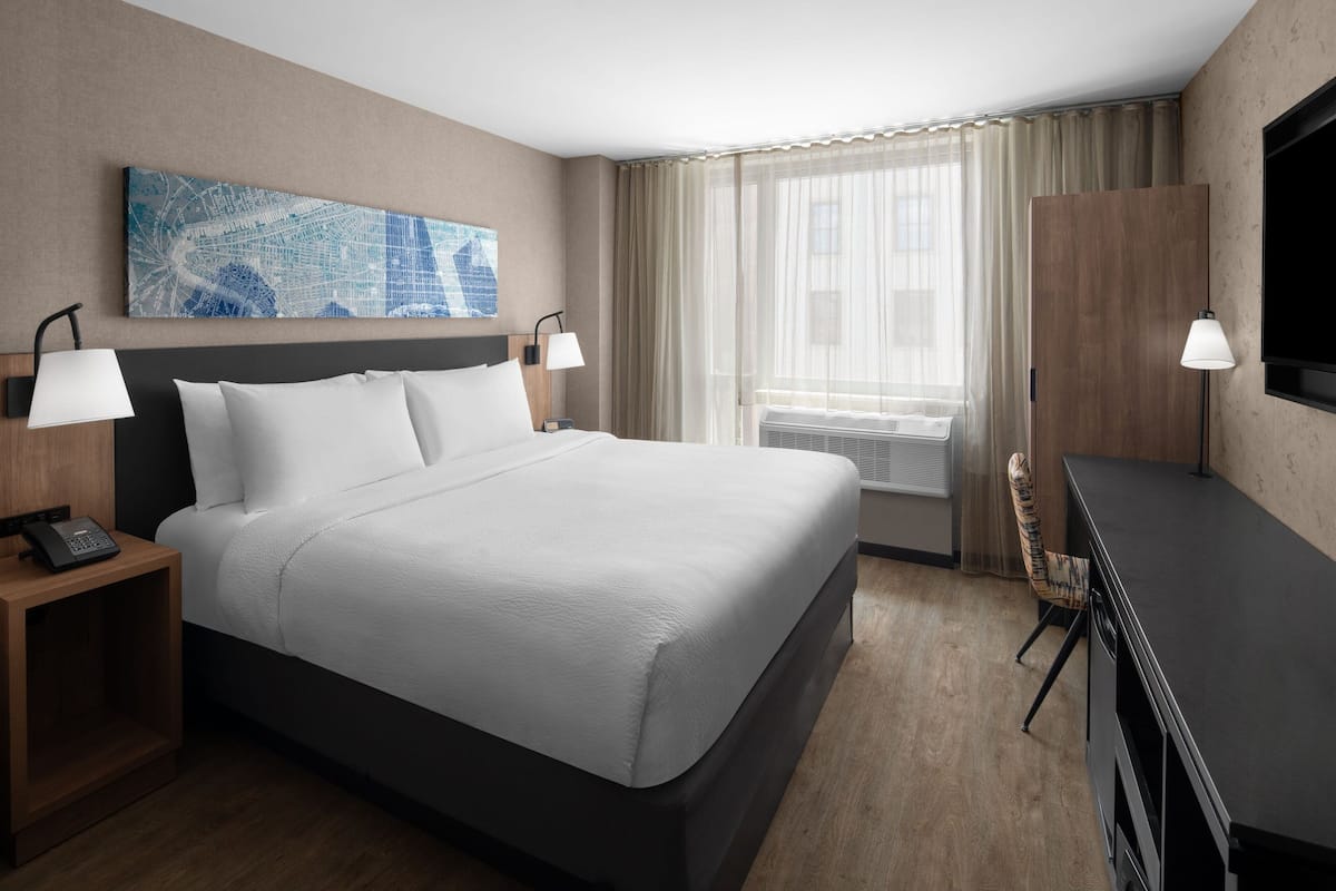 Ein Hotelzimmer mit einem großen Bett, zwei Nachttischen mit Lampen, einem Schreibtisch, einem Stuhl, einem Flachbildfernseher, einem Fenster mit Vorhängen und einem Stadtbild an der Wand.