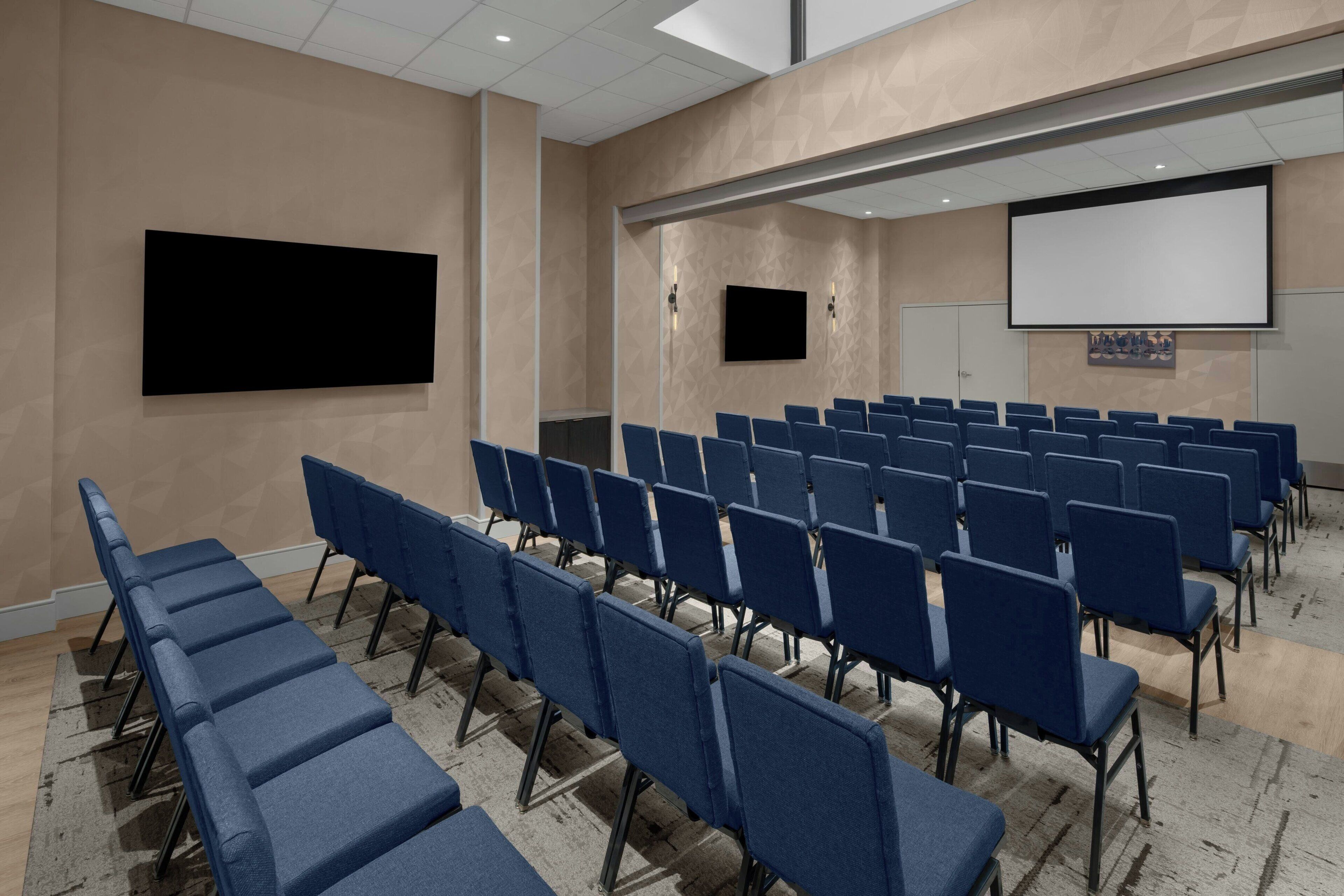 Un salón de conferencias con filas de sillas azules, una pantalla grande y una pantalla de proyección.