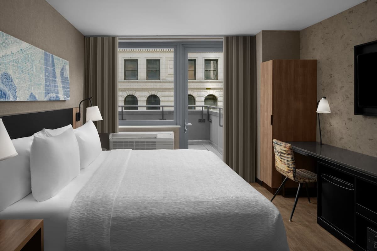 Ein Hotelzimmer mit einem großen Bett, einem Schreibtisch mit Stuhl, einem Fernseher und Blick auf ein Gebäude durch eine Glastür.