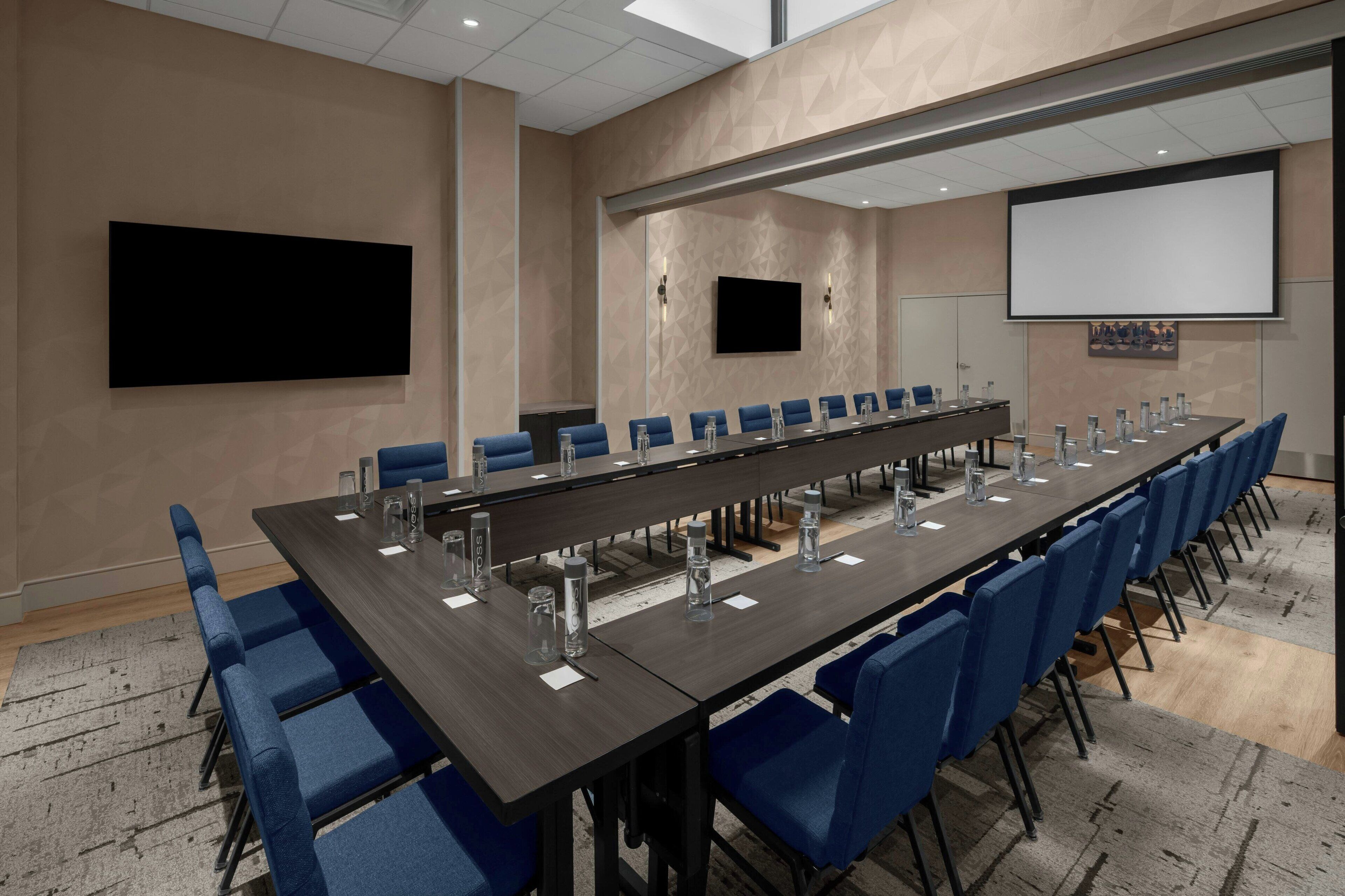 Una sala de conferencias con una larga mesa, sillas azules y una pantalla para proyector.