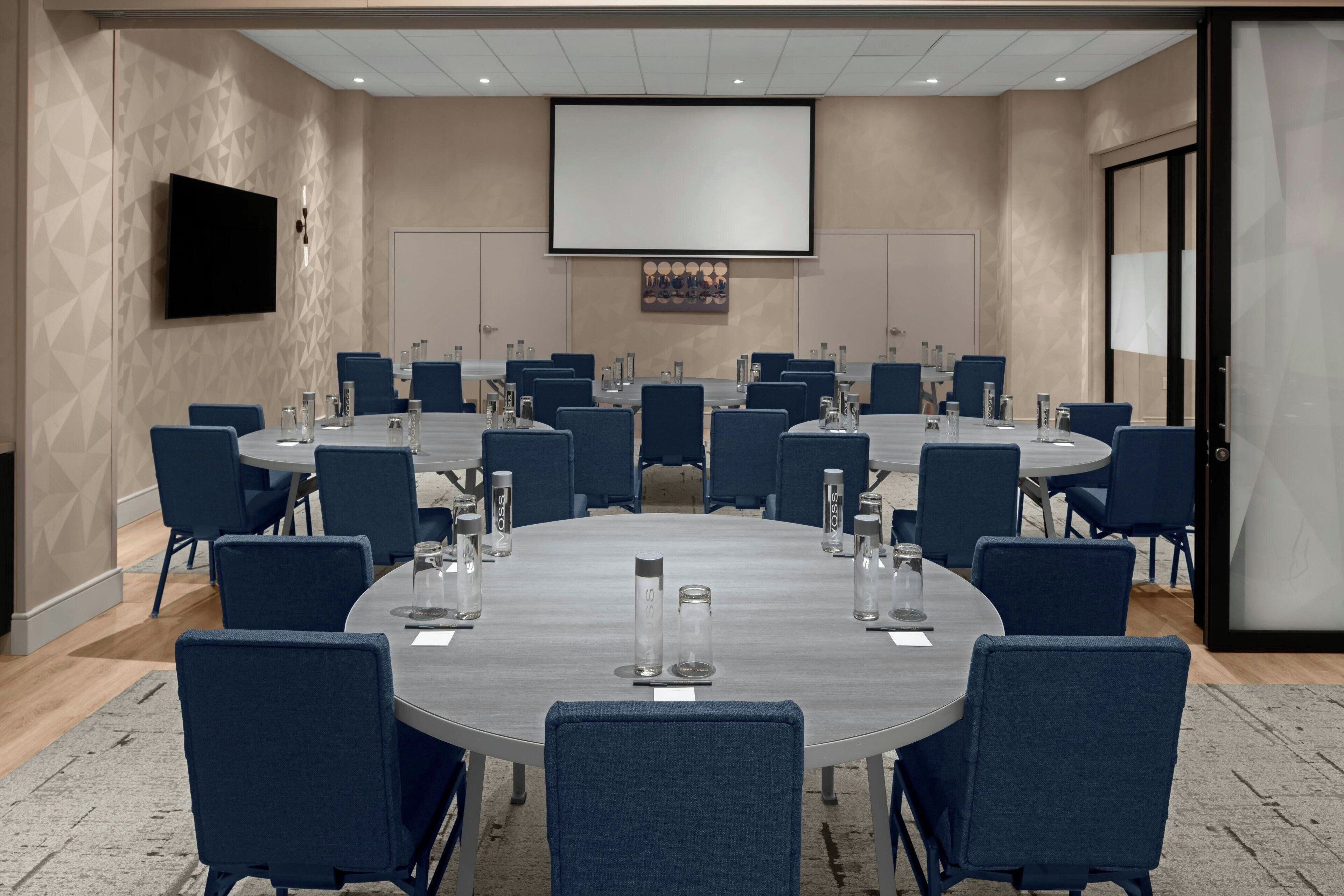 Un salón de conferencias con mesas redondas, sillas y una pantalla grande.