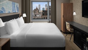 A hotel room with a large bed, a desk, a chair, a TV, and a window with a city view.