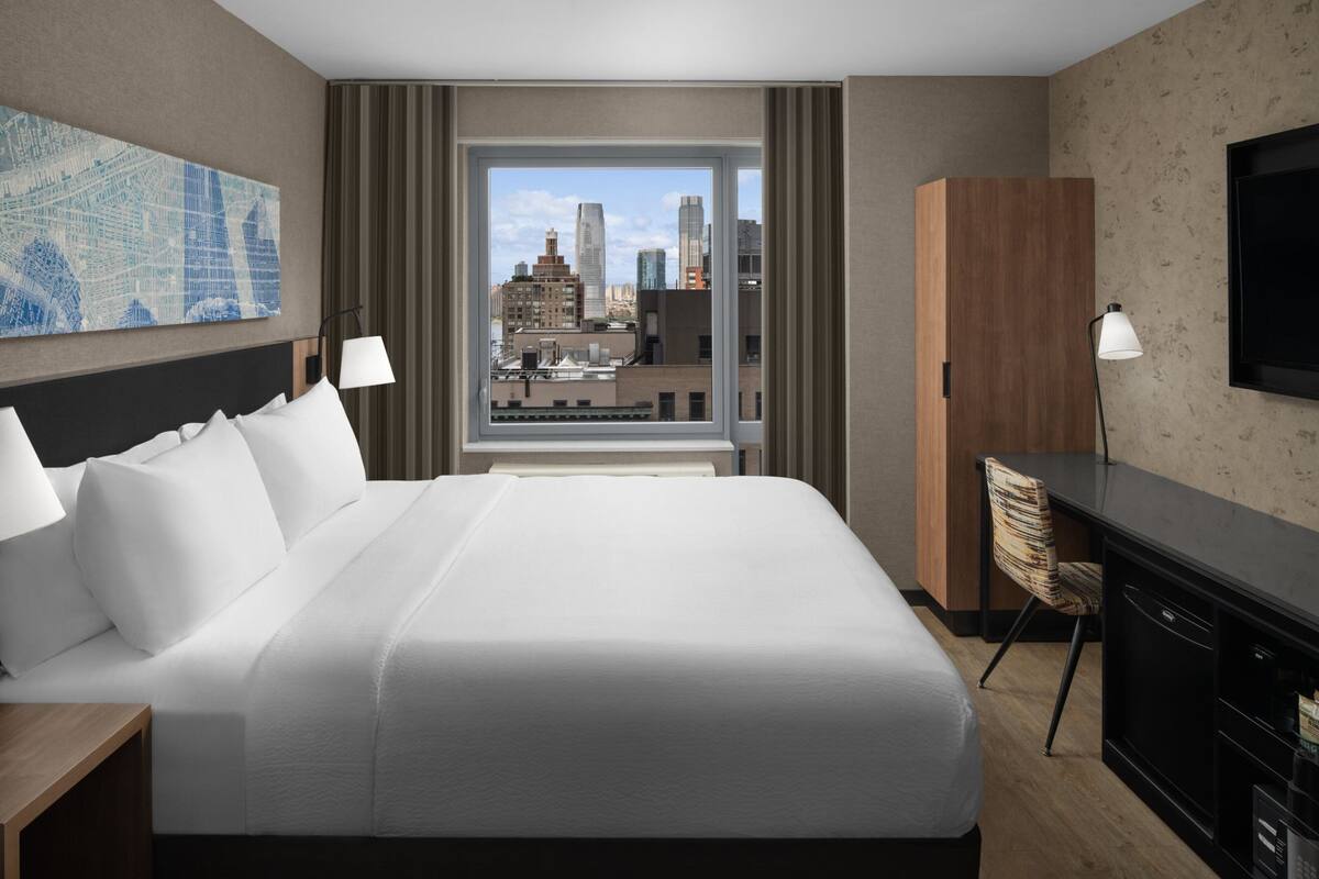 Ein Hotelzimmer mit einem großen Bett, einem Schreibtisch, einem Stuhl, einem Fernseher und einem Fenster mit Blick auf die Stadt.
