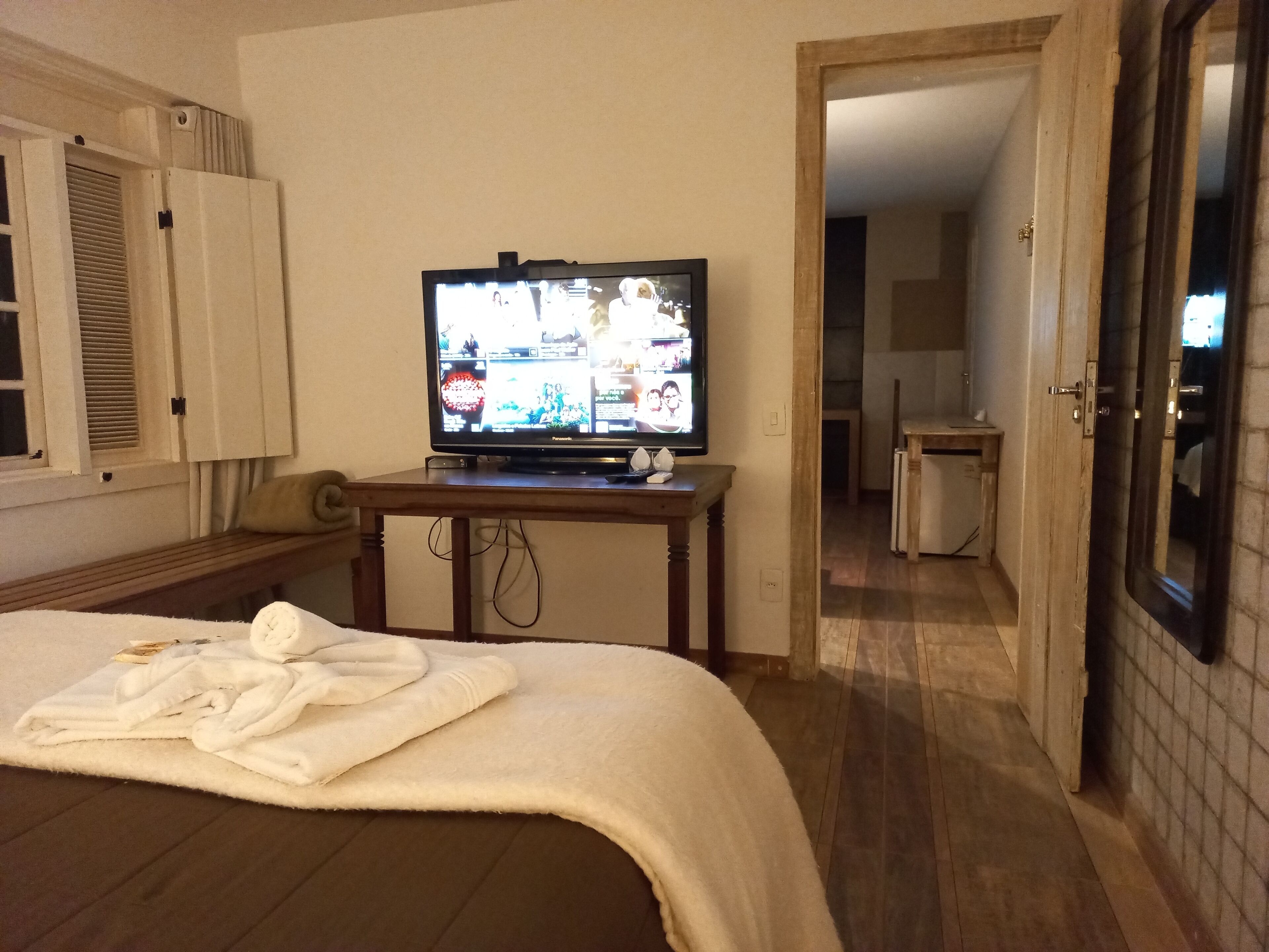 Habitación de hotel con cama, un televisor sobre una mesita de madera, baño visible a través de una puerta abierta y una ventana con persianas.