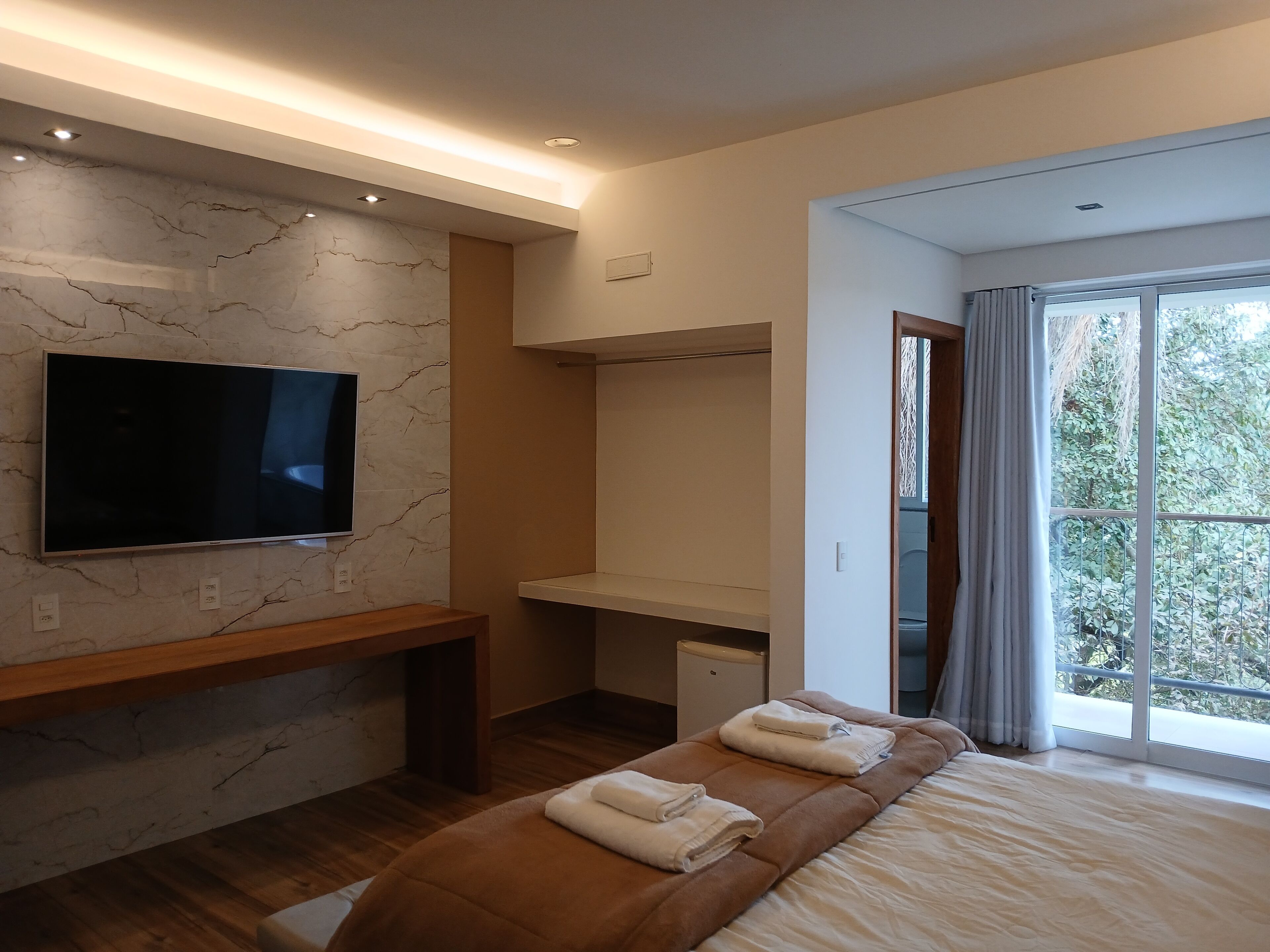 Un dormitorio moderno con una televisión de pantalla plana montada en una pared de mármol, un banco de madera, un escritorio y una cama grande con toallas.