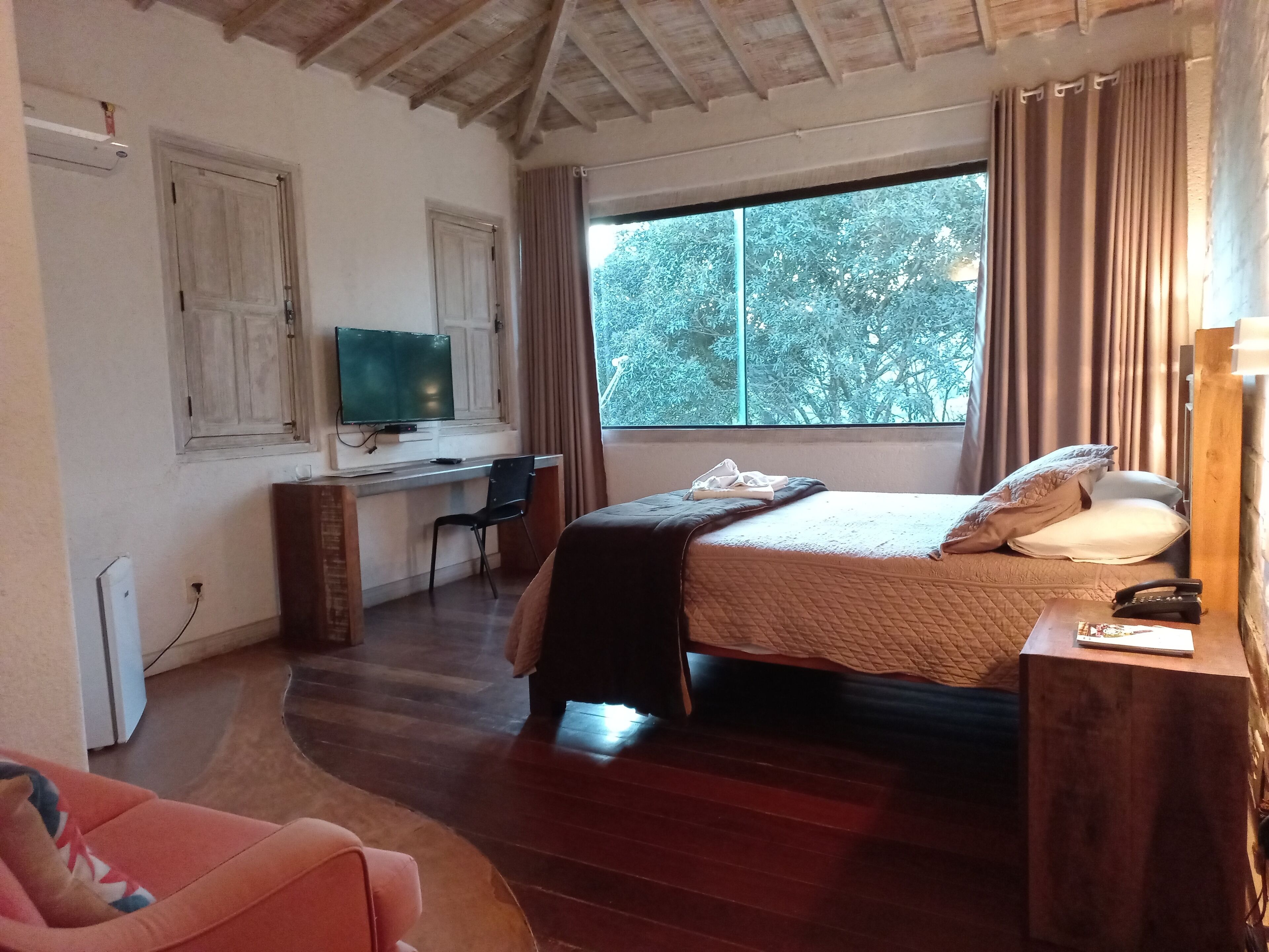 A bedroom with a bed, a desk with a computer, a chair, a TV, and a window with curtains.