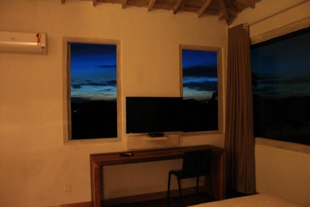 A room with a bed, a desk with a chair, a TV, and two windows with a view of the sky.