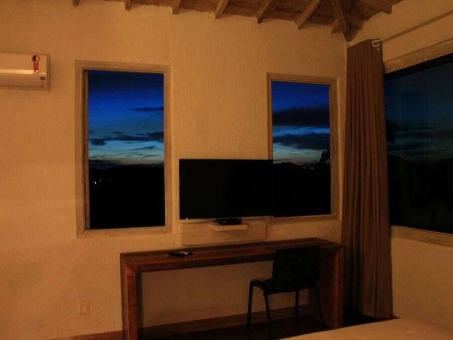 A room with a bed, a desk with a chair, a TV, and two windows with a view of the sky.