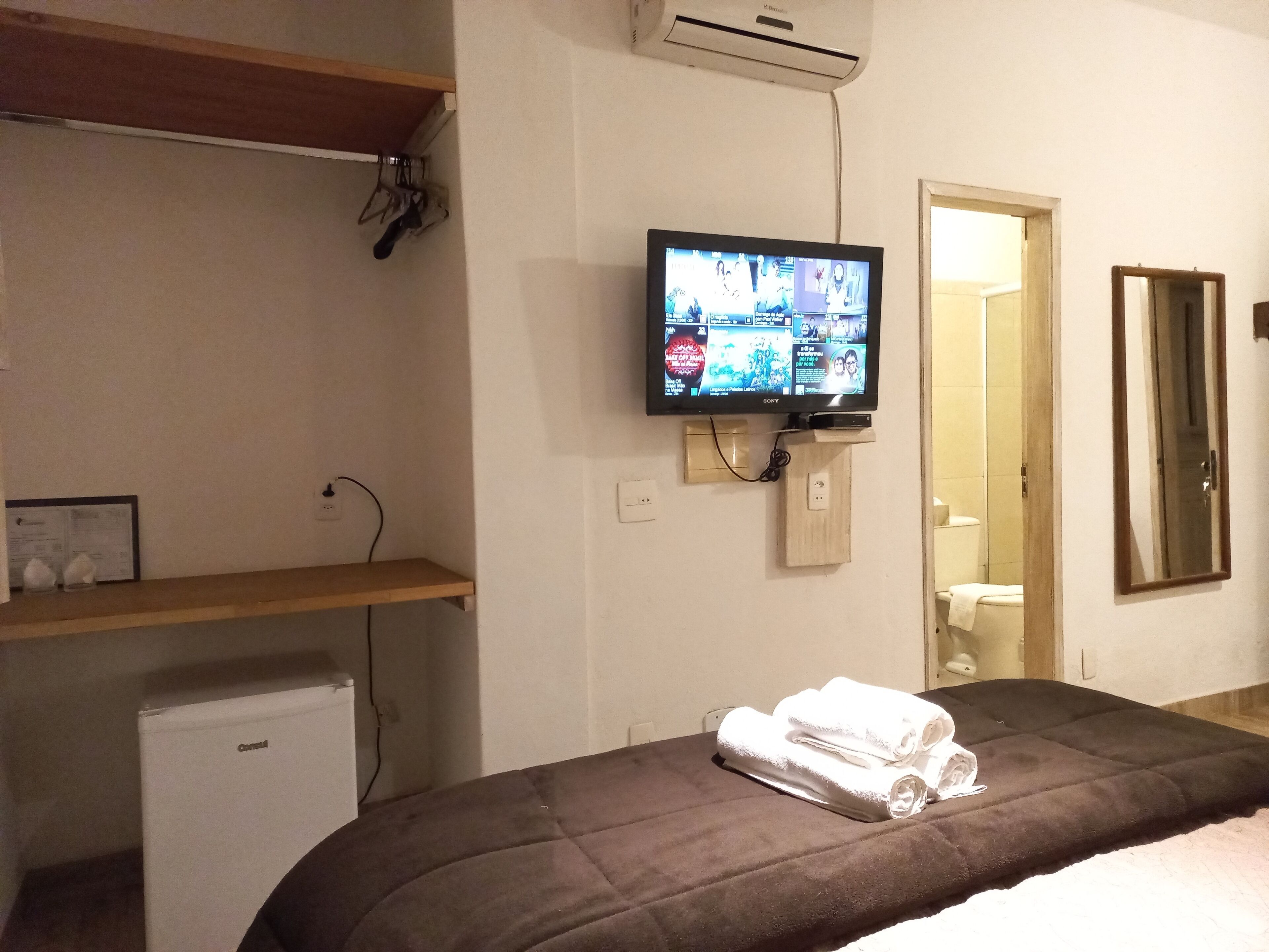 A hotel room with a bed, a TV mounted on the wall, a small fridge, and a wooden shelf.