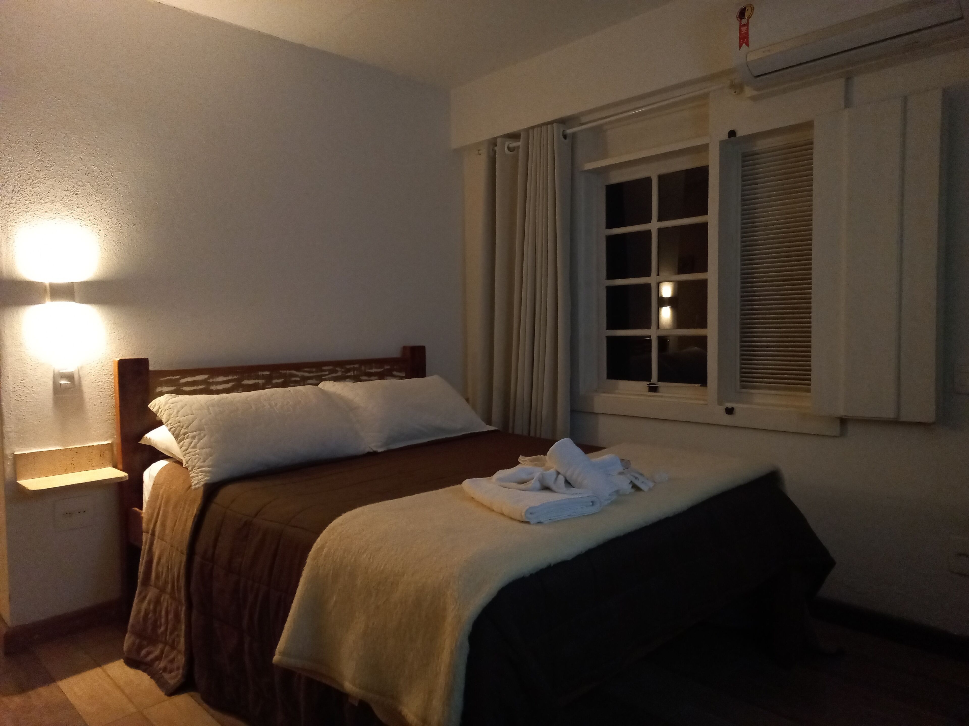 A bedroom with a bed, a nightstand, a wall-mounted light, a window with blinds, and a thermostat.