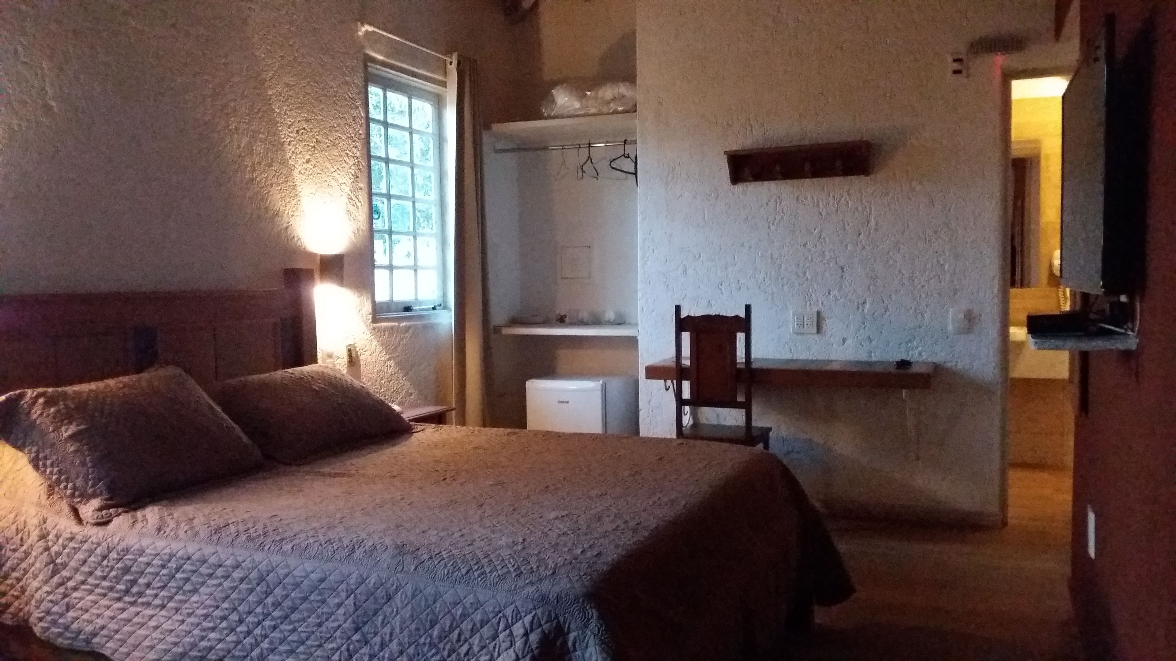 Un dormitorio con una cama, una silla, una mesita, una ventana y un televisor instalado en la pared.