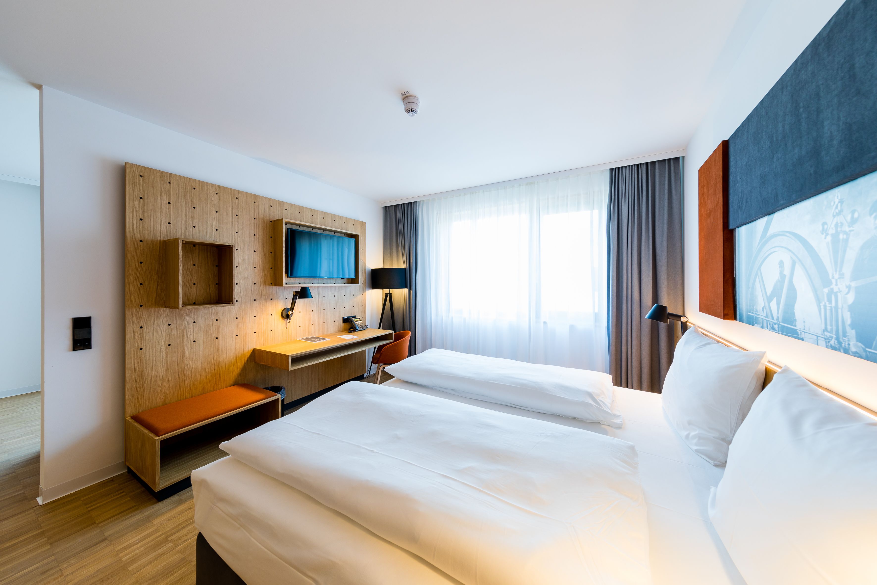 A hotel room with two beds, a desk, a TV, and a large window with curtains.