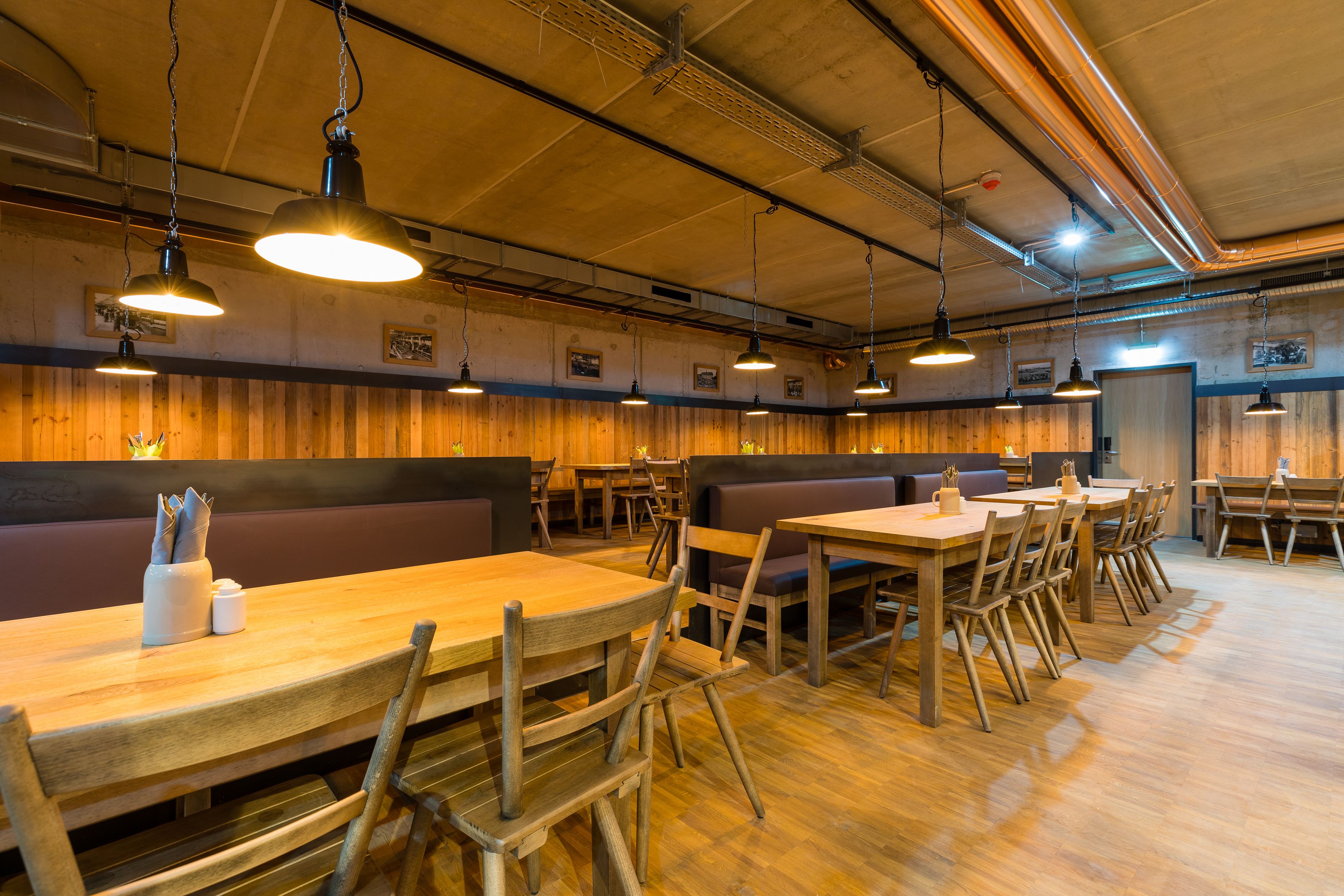 A restaurant interior with wooden tables and benches, pendant lighting, and exposed ceiling pipes.