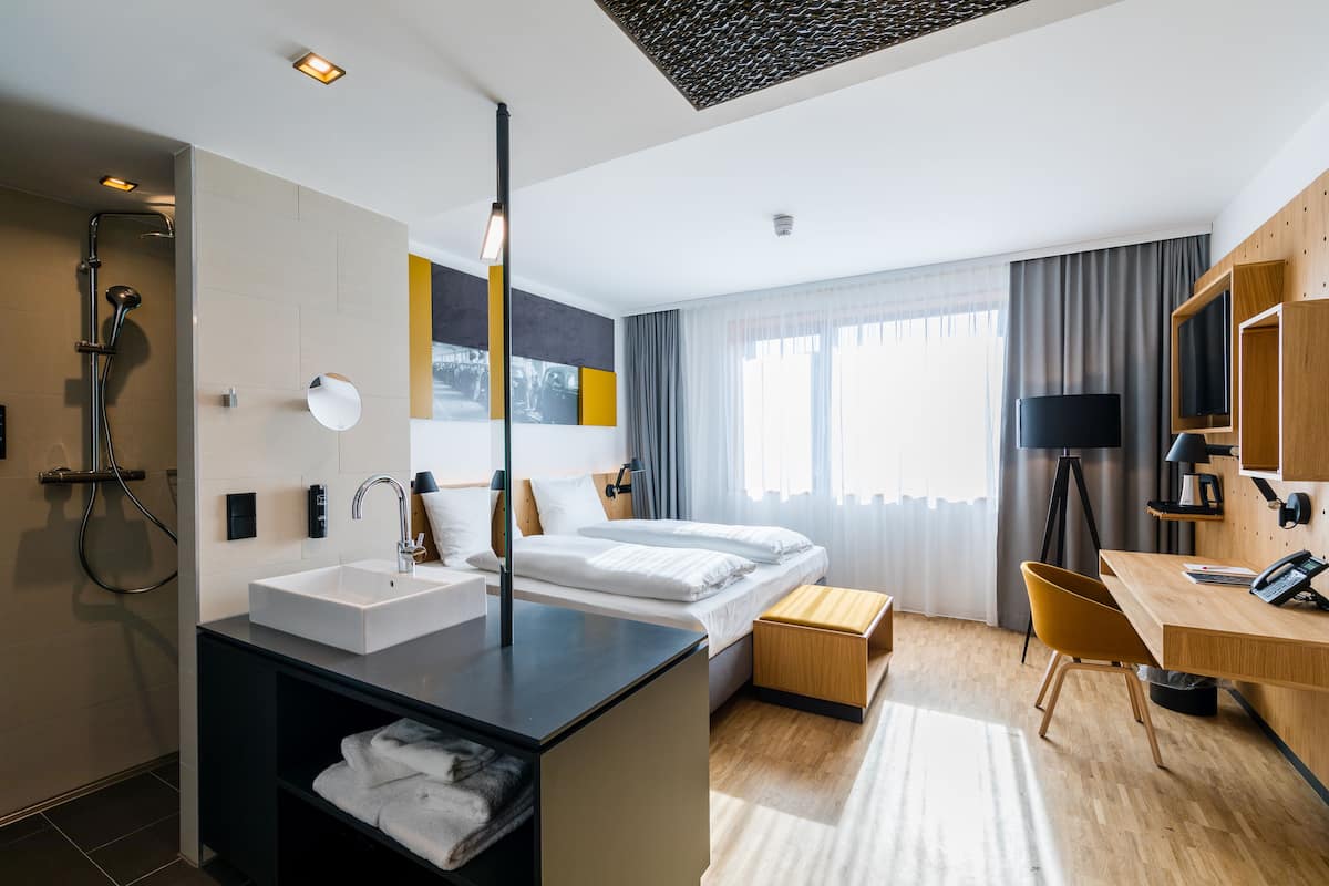 Una habitación de hotel moderna con una cama grande, un escritorio, una silla, una lámpara de escritorio, un televisor, una ducha y un lavamanos.