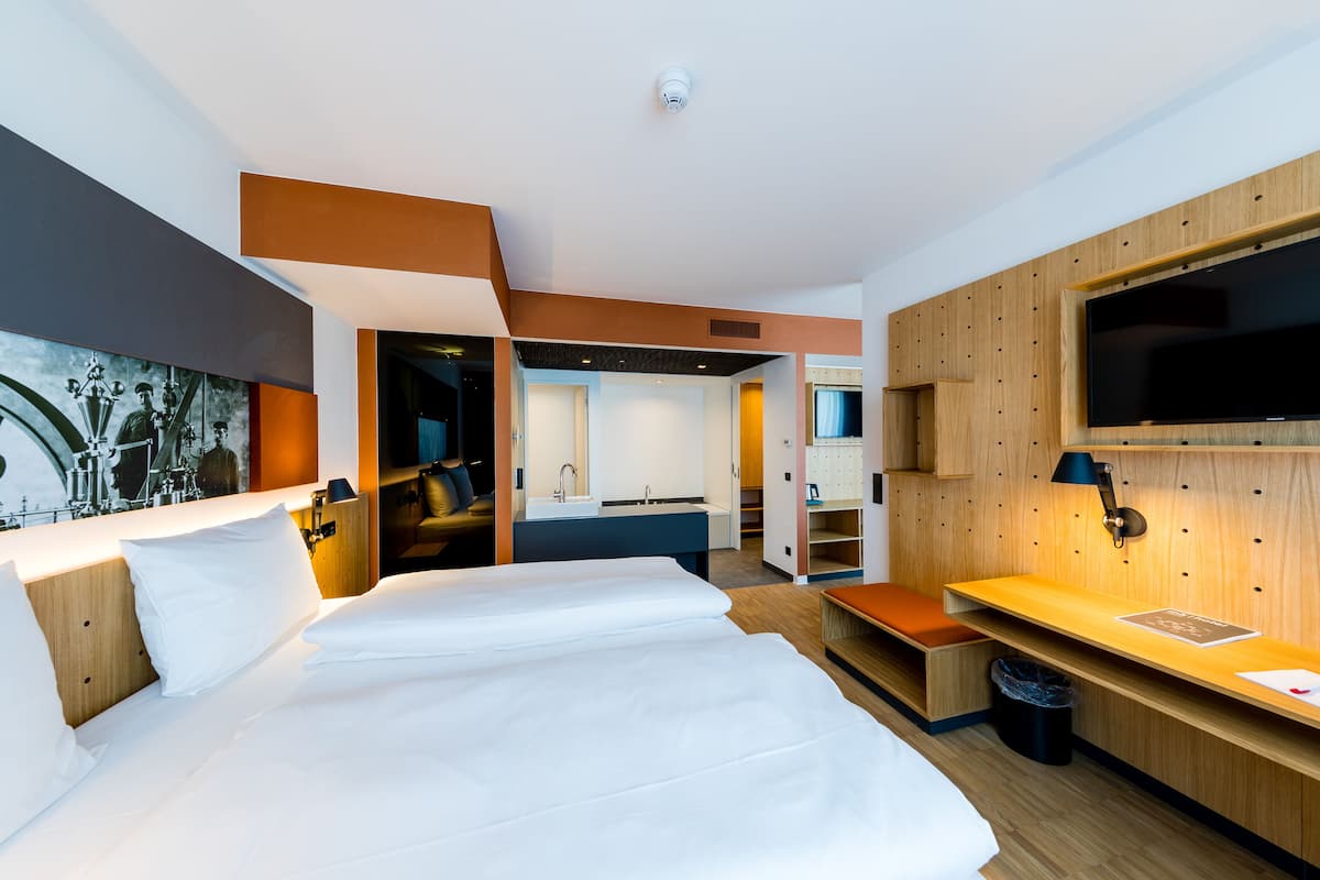 Una habitación de hotel moderna con una cama grande, un escritorio, un televisor y un baño visible a través de una puerta abierta.